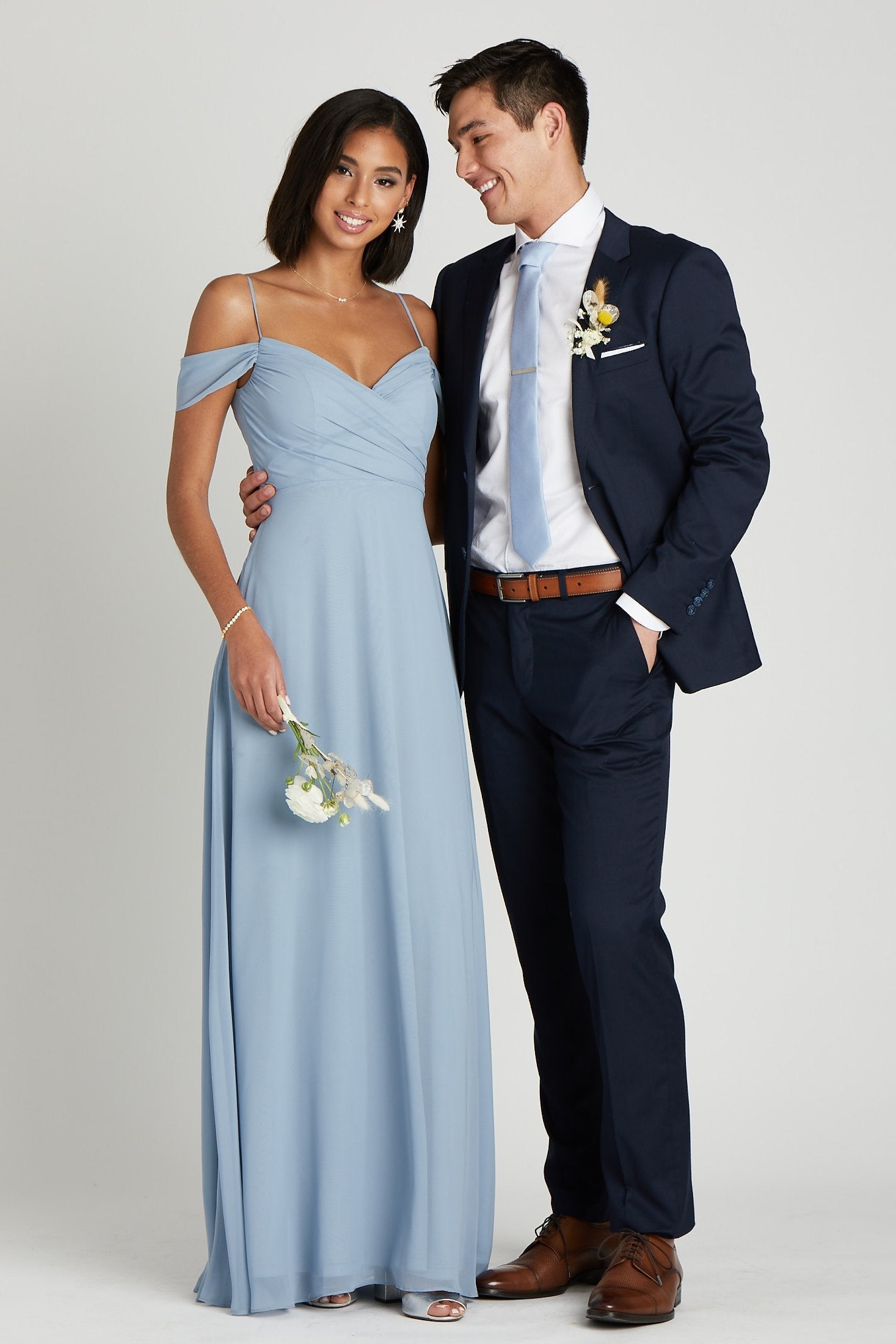 Front view of two models in a side embrace at the waist. One model with a slender physique and a medium skin tone wears the Simon Necktie in dusty blue with a white collared button down shirt and the Navy Blue Suit by Suitshop. The second model with a slender figure and a medium skin tone coordinates their look with a floor length dress in dusty blue.