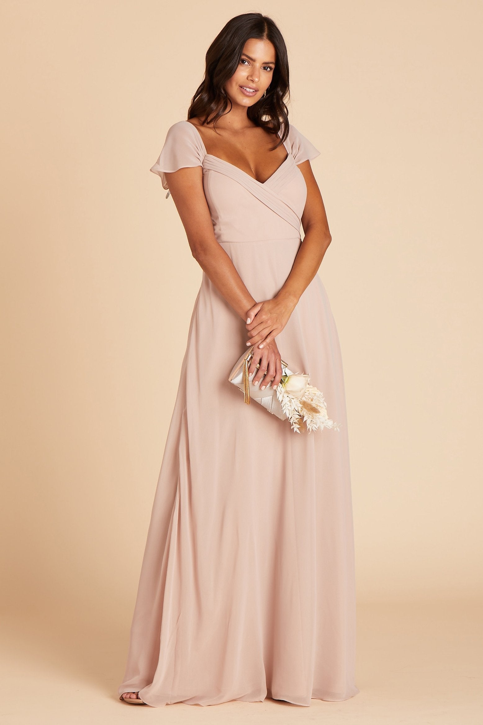 Front view of the Spence Convertible Dress in taupe chiffon shows the model holding a dried flower ivory bouquet. The bodice front shows a smooth fit with cleavage showing due to the built-in boning and bra cups. The dress sleeves rest on their shoulders and flow onto their arm. 