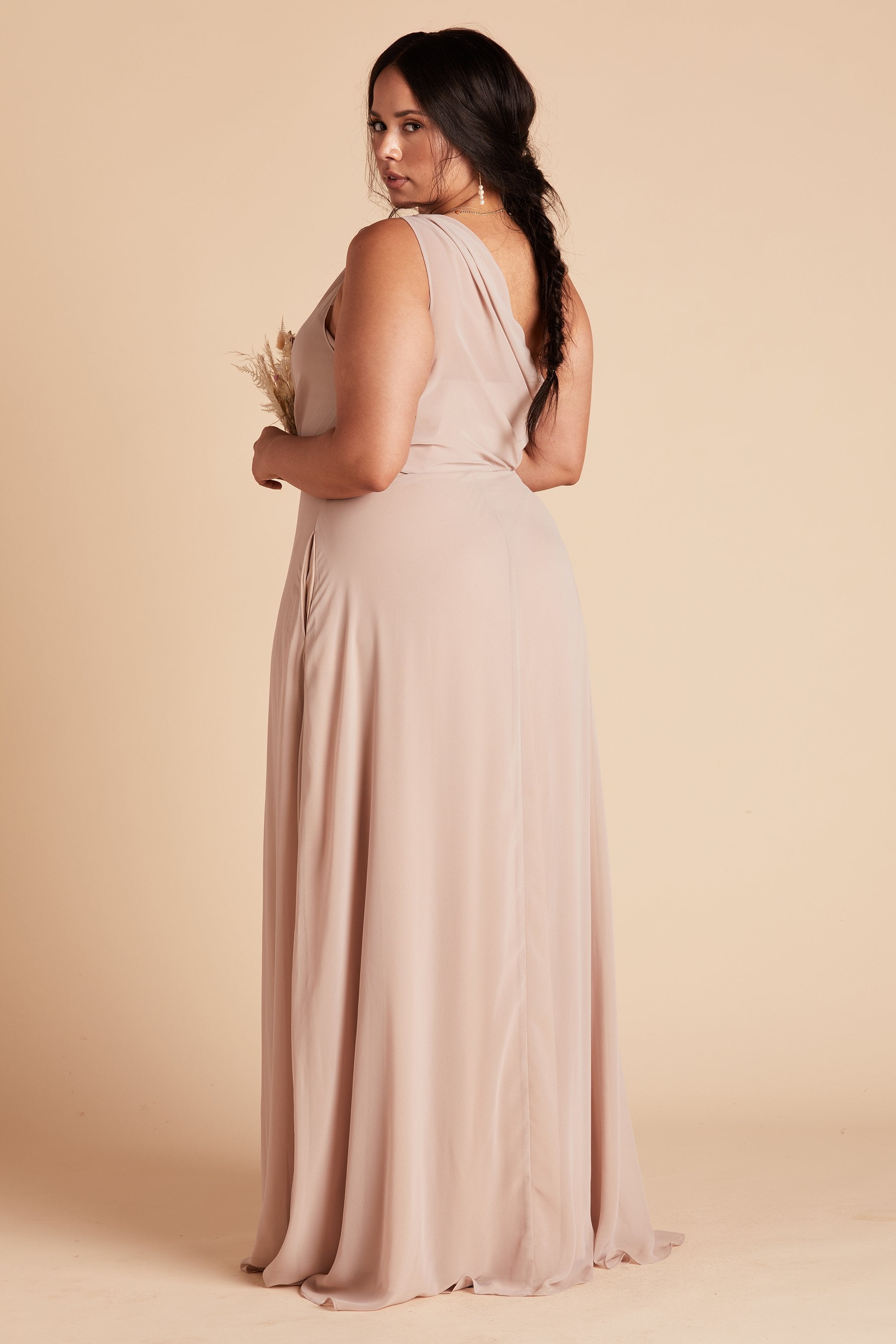 Back and side view of the Kira Dress Curve in taupe chiffon shows a full-figured model with medium skin wearing a sheer, softly pleating chiffon gathered at the bodice shoulder and draping across the back. The left side seam has an inset pocket. 