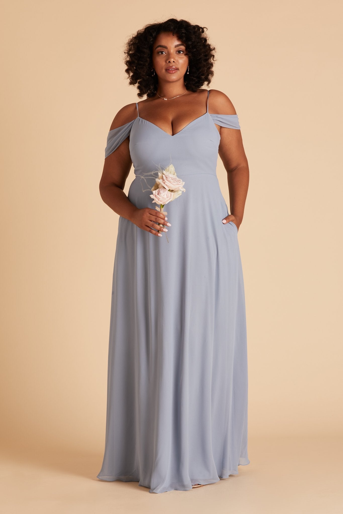 Front view of the floor-length Devin Convertible Plus Size Bridesmaid Dress in dusty blue chiffon worn by a curvy model with a medium dark skin tone. The dress detachable sleeves drape gracefully over the model’s upper arms.