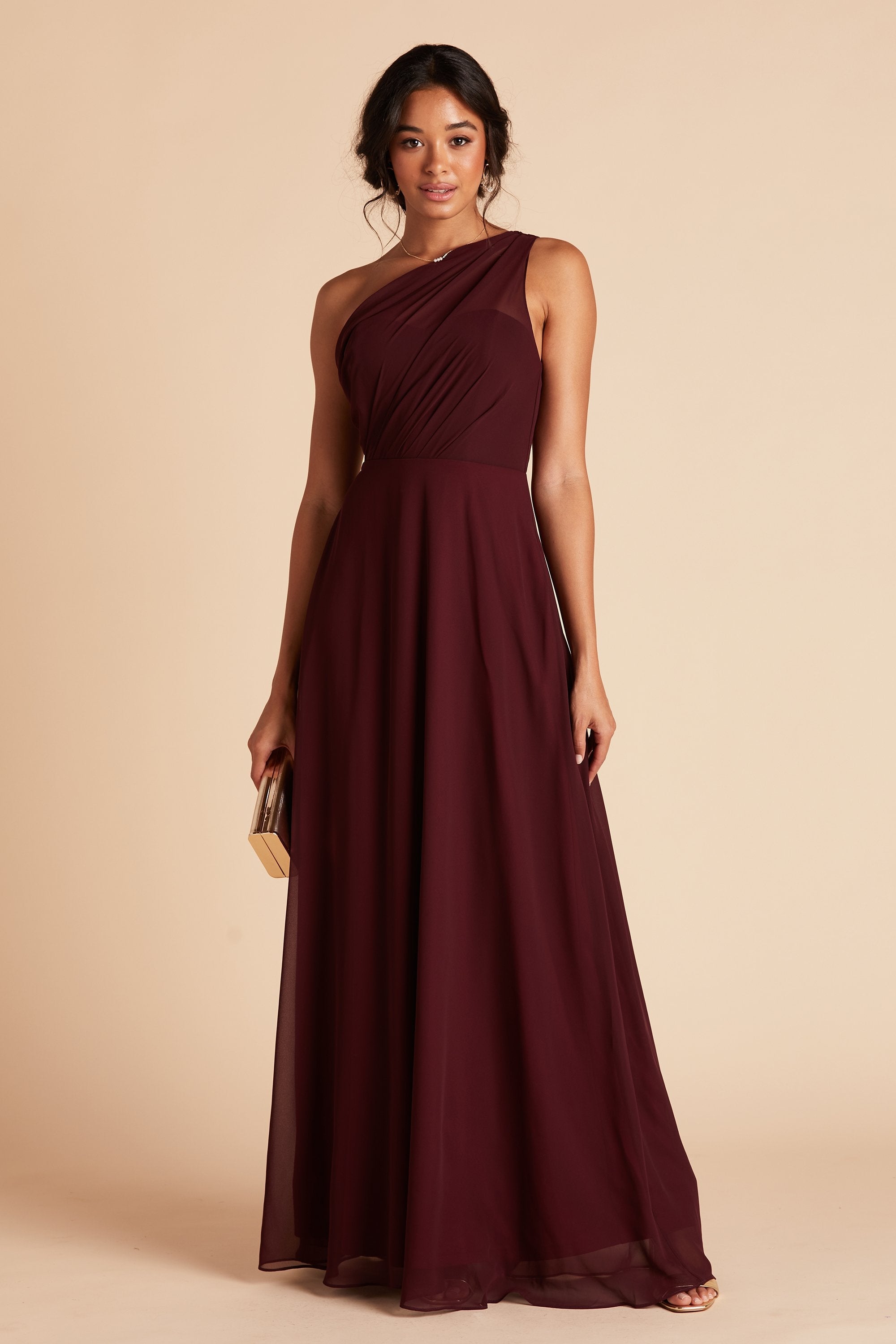 Front view of the Kira Dress in cabernet chiffon without the optional slit shows a slender model with a medium skin tone holding a clutch purse at her side. 