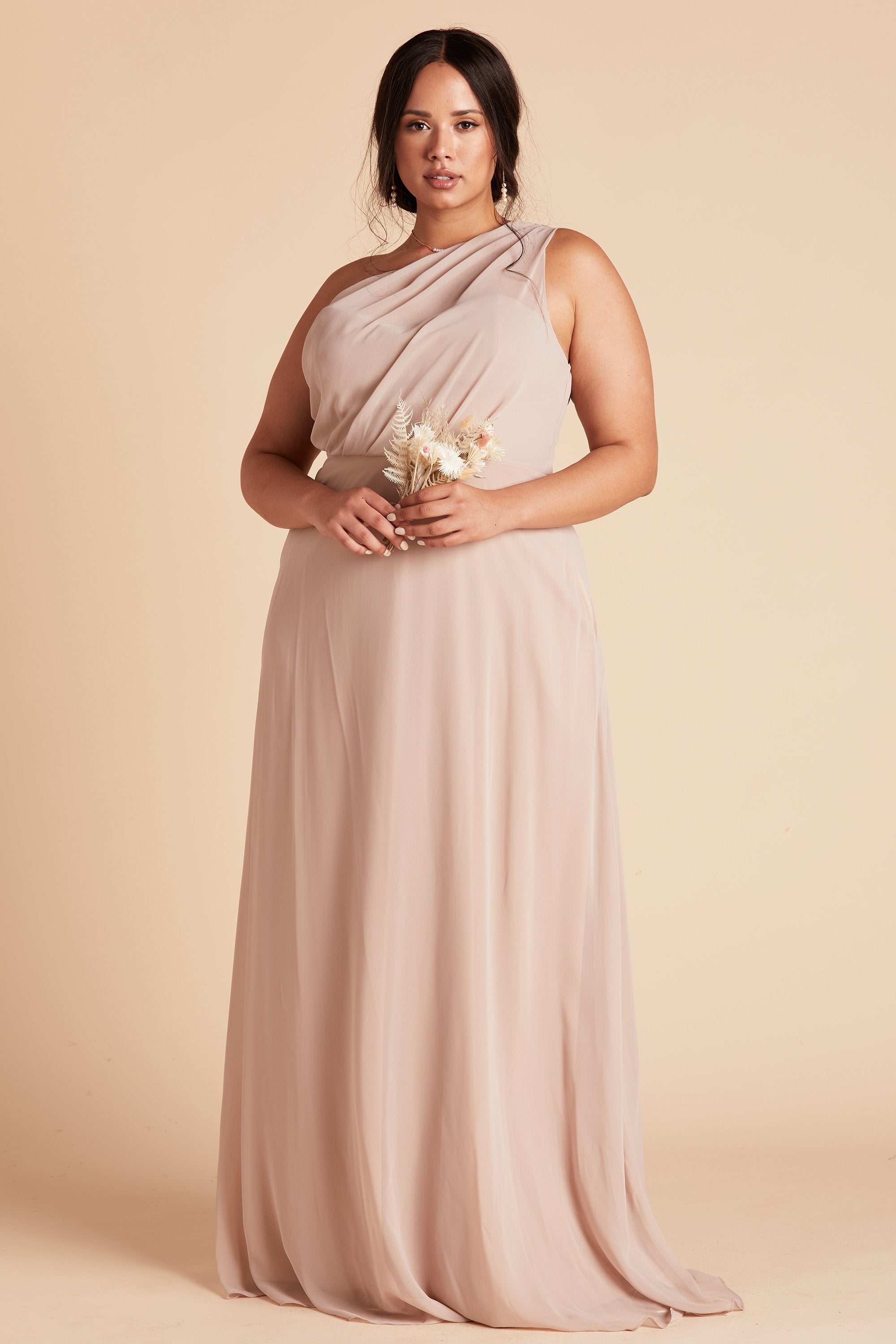 Front view of the Kira Dress Curve in taupe chiffon without the optional slit shows a full-figured model with a medium skin tone wearing an asymmetrical one-shoulder, full-length dress. Soft pleating gathers at the left shoulder of the bodice with a smooth fit at the waist as the A-line dress flows to the floor.