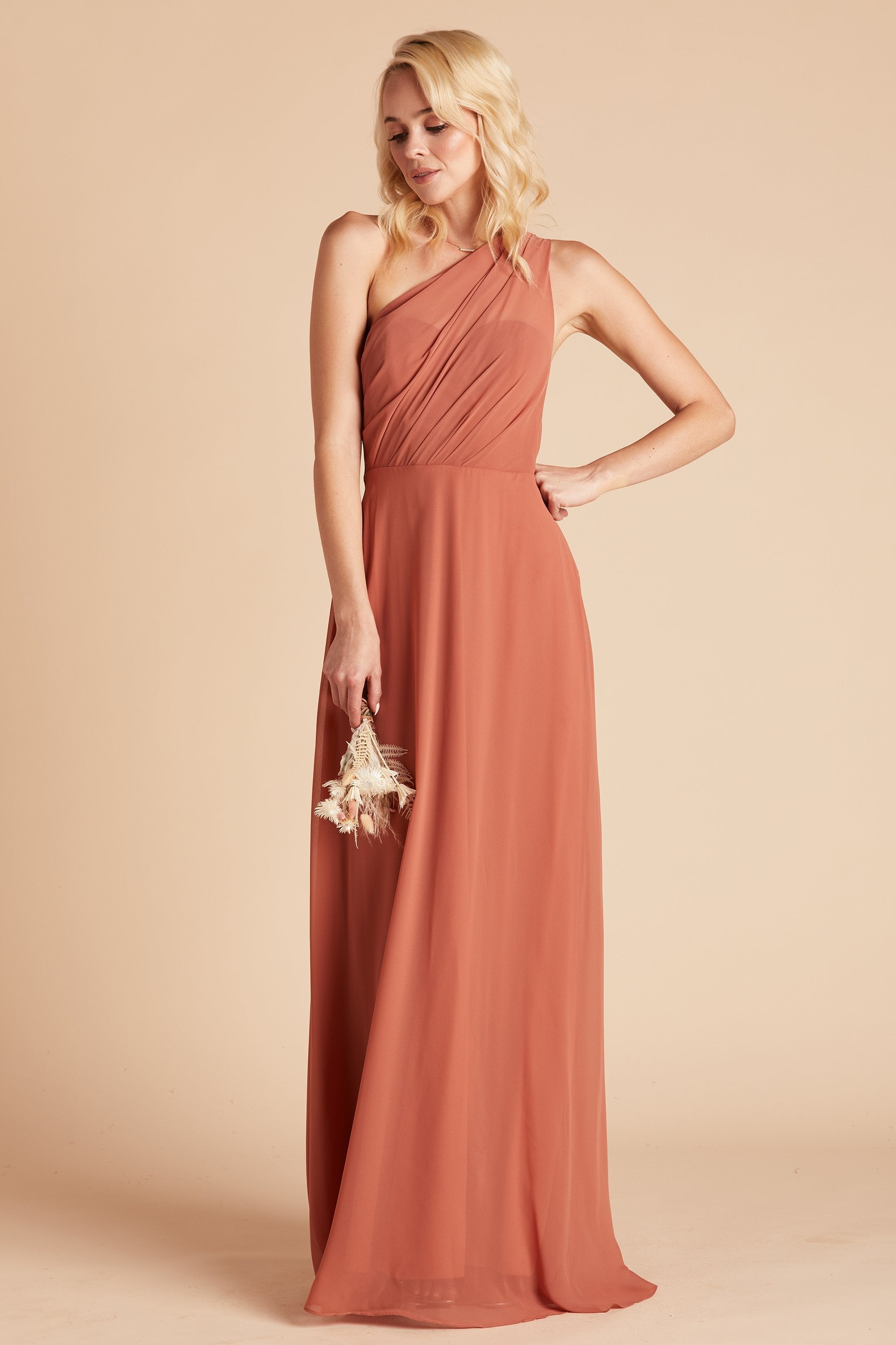 Front view of the Kira Dress in terracotta chiffon without the optional slit shows a slender model with a light skin tone wearing an asymmetrical one-shoulder, full-length dress. Soft pleating gathers at the left shoulder of the bodice with a smooth fit at the waist as the dress skirt with a slight A-line silhouette flows to the floor.