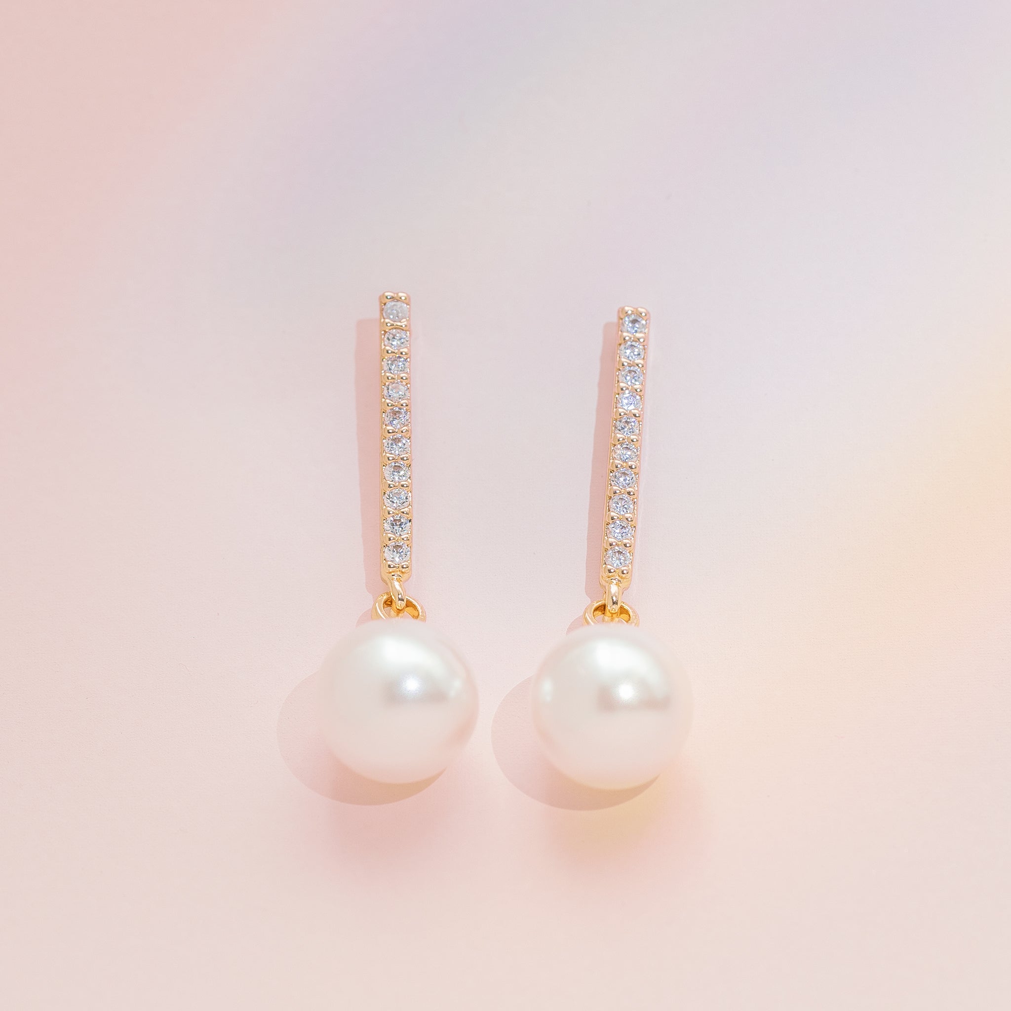 Close-up view of Cincinnati Pearl Drop Earrings. Each earring features a row of ten cubic zirconia in gold-colored brass above a dangling plastic pearl.