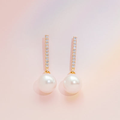 Close-up view of Cincinnati Pearl Drop Earrings. Each earring features a row of ten cubic zirconia in gold-colored brass above a dangling plastic pearl.