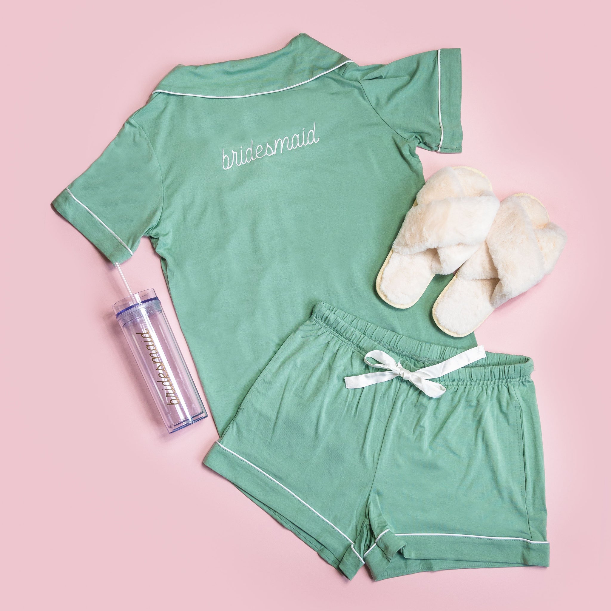 From above, we see the Johnny Pajama Set in sea glass laid out flat, with a pair of Fuzzy Slippers in white by Birdy Grey and a clear water bottle. The pajama top and the water bottle are personalized with the text “Bridesmaid.”