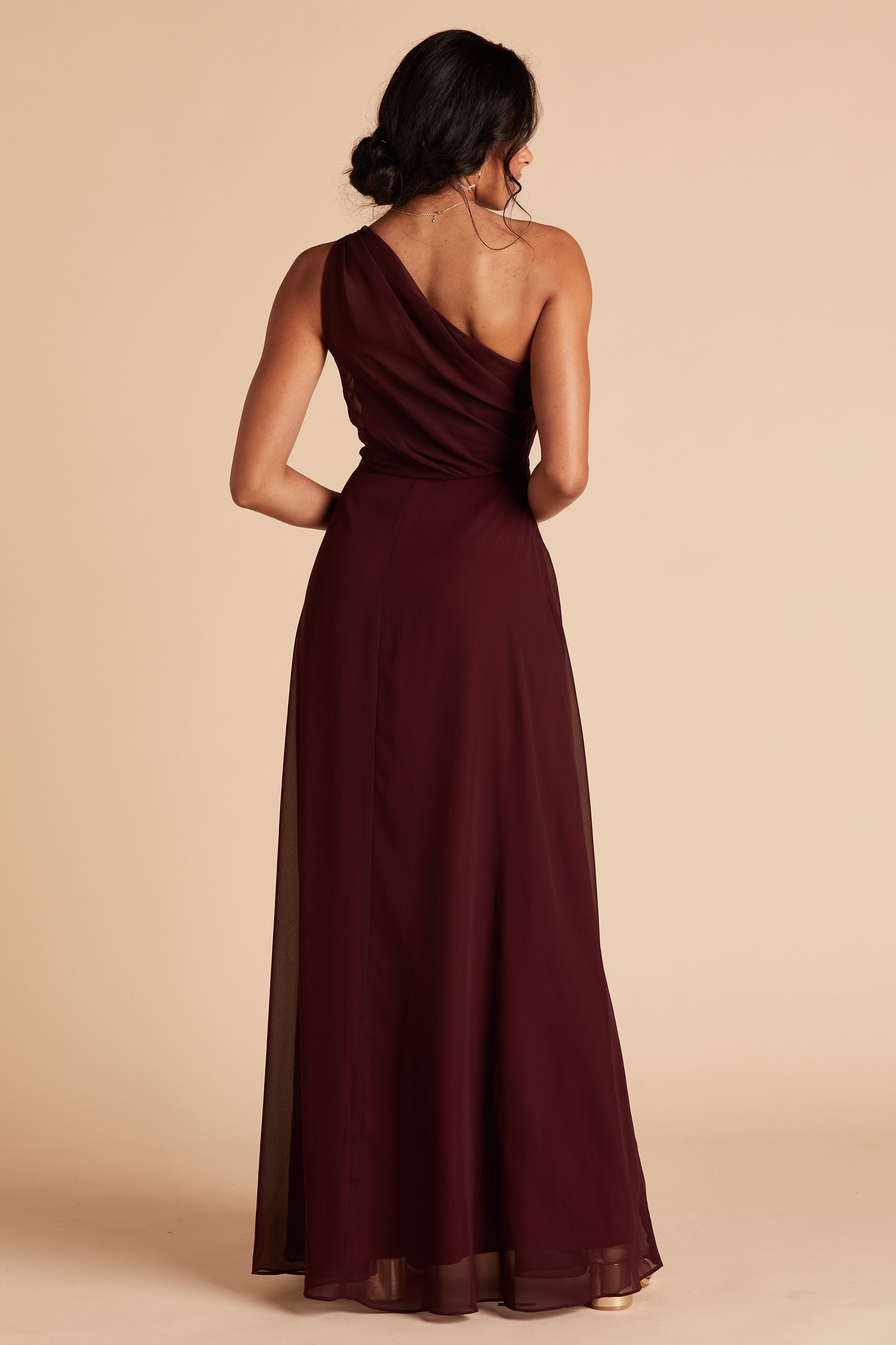 Back view of the Kira Dress in cabernet chiffon shows a slender model with a medium skin tone. The back of the dress has sheer chiffon fabric which creates a pleated Grecian one-shoulder neckline and bodice.