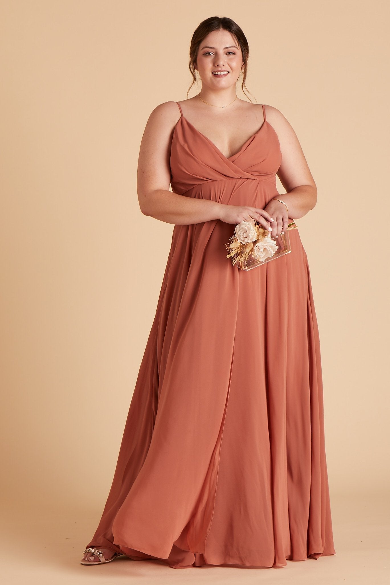 Front view of the Kaia Dress Curve in terracotta chiffon shows the model holding a small dried flower bouquet and the Clear Clutch with Clear Beads purse. The V-neck and wide pleated bodice overlaps across the chest and the dress skirt front falls gently at the sides.