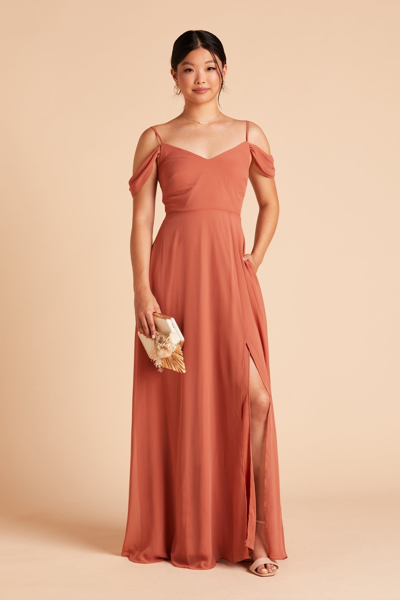 Front view of the floor-length Devin Convertible Bridesmaid Dress in terracotta chiffon worn by a slender model with a light skin tone. The model rests her left hand in the hidden side pocket.