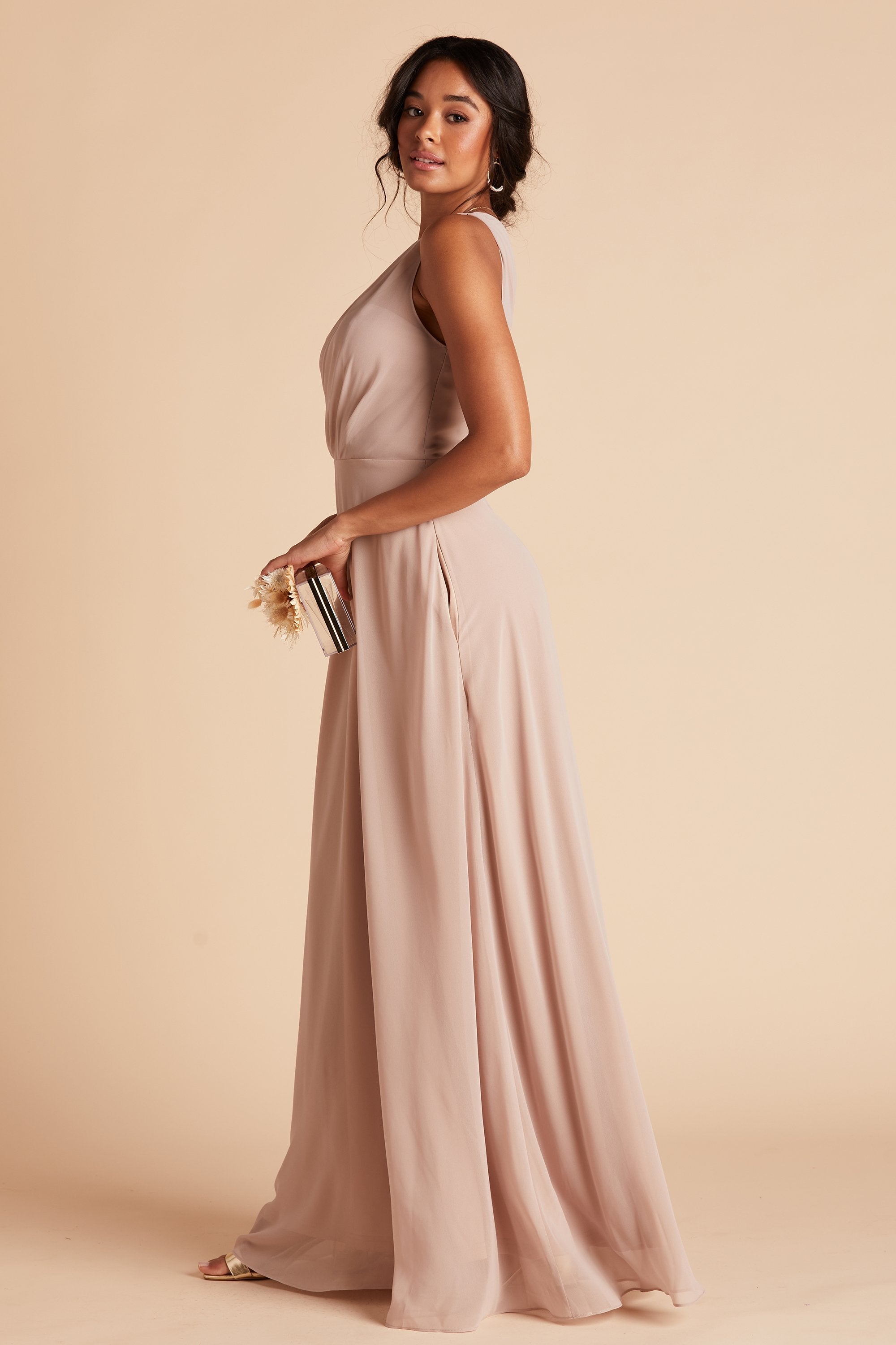 Side view of the Kira Dress in taupe chiffon shows a slender model with a medium skin tone with side seam pocket showing. They are holding the Clear Clutch with Clear Beads purse.