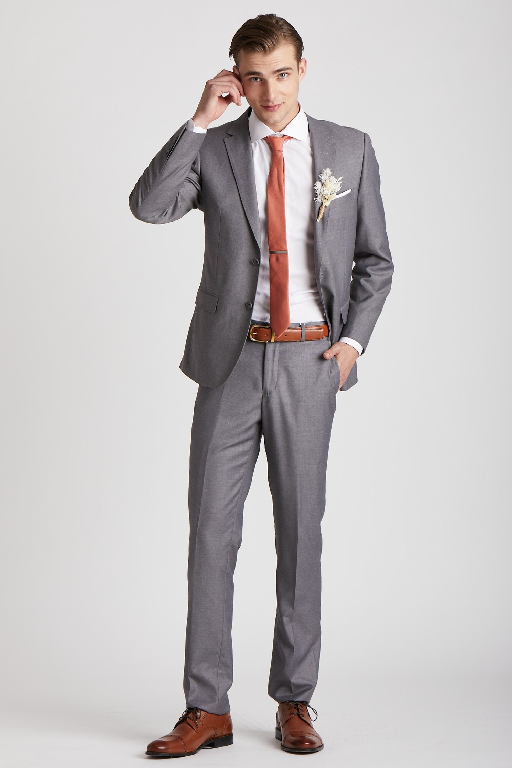 Front view of a model wearing the Simon Necktie in terracotta coordinated with a medium grey suit with white collared shirt, medium brown shoes and belt, white pocket square, and a dried flower boutonniere in ivory. 