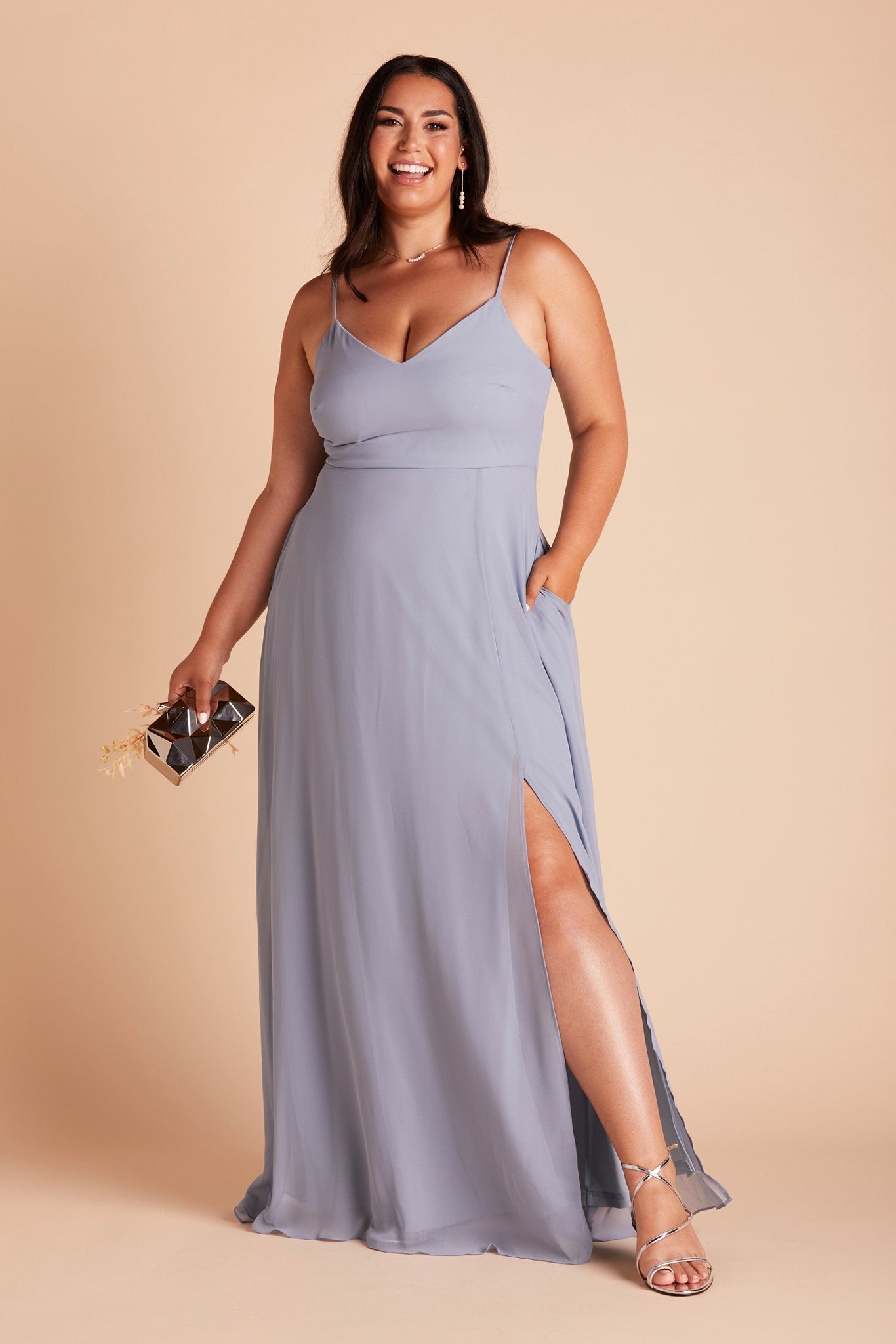 Front view of the floor-length Devin Convertible Plus Size Bridesmaid Dress in dusty blue chiffon worn by a curvy model with a light skin tone. The model rests her left hand in the hidden side pocket.