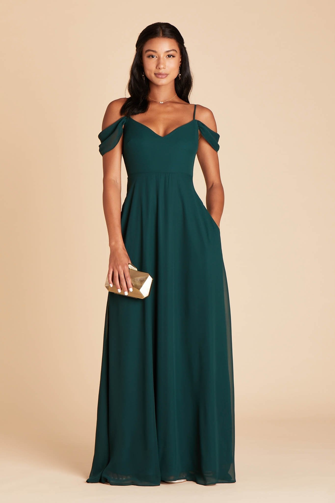 Front view of the floor-length Devin Convertible Bridesmaid Dress in emerald chiffon by Birdy Grey with a V-neck front and draping detachable sleeves. The model rests her left hand in the hidden side pocket.