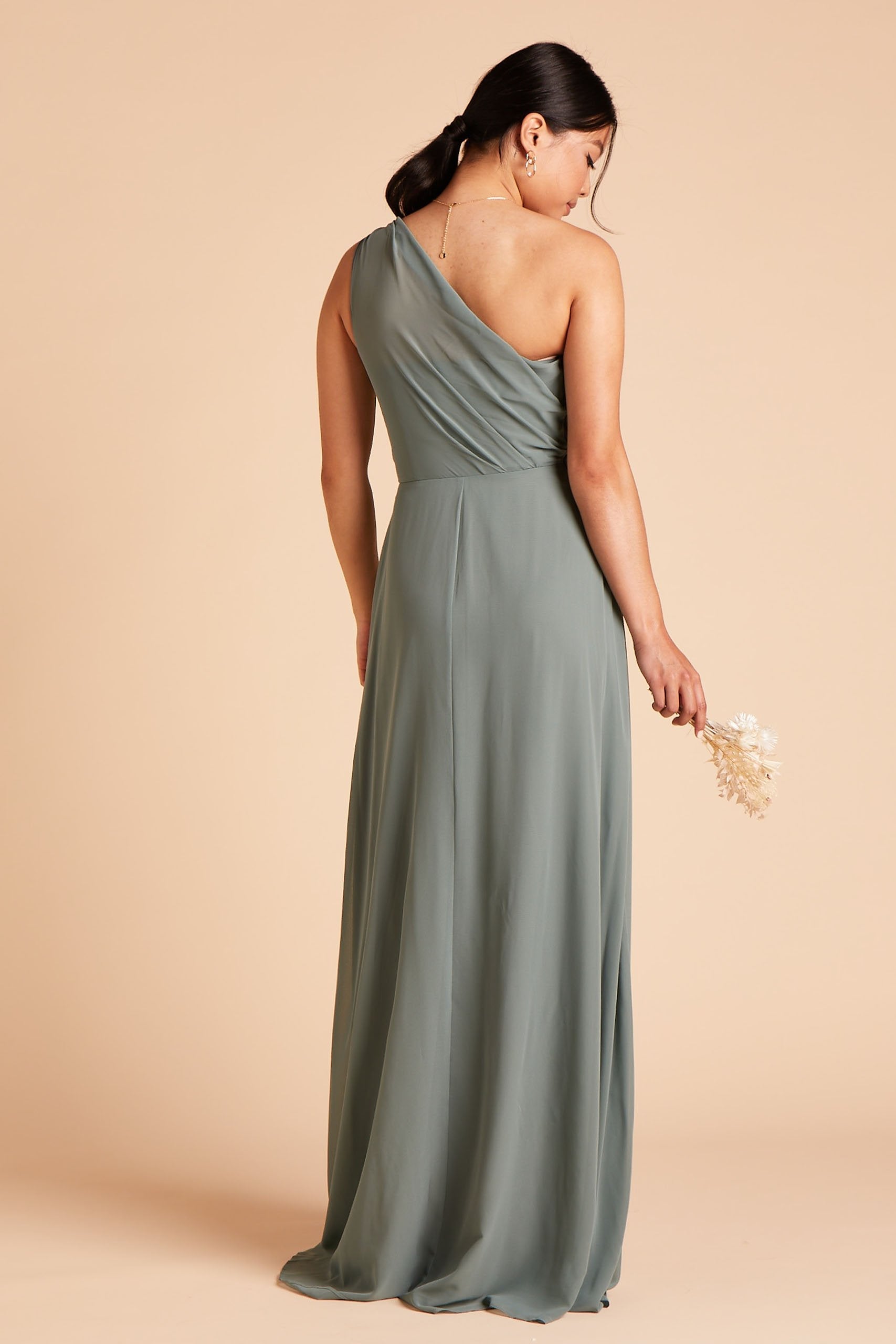 Back view of the Kira Dress in sea glass chiffon shows a model wearing sheer, softly pleating chiffon gathered at the bodice shoulder and draping across the back to the side seam as the dress flows to the floor. 