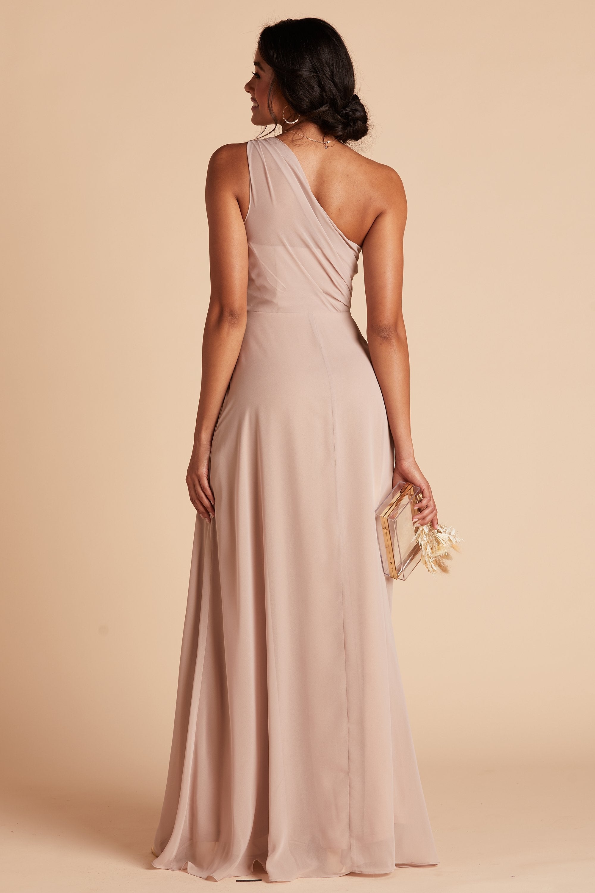 Back view of the Kira Dress in taupe chiffon shows a slender model with medium skin wearing sheer, softly pleating chiffon gathered at the bodice shoulder and draping across the back to the side seam as the dress flows to the floor. 