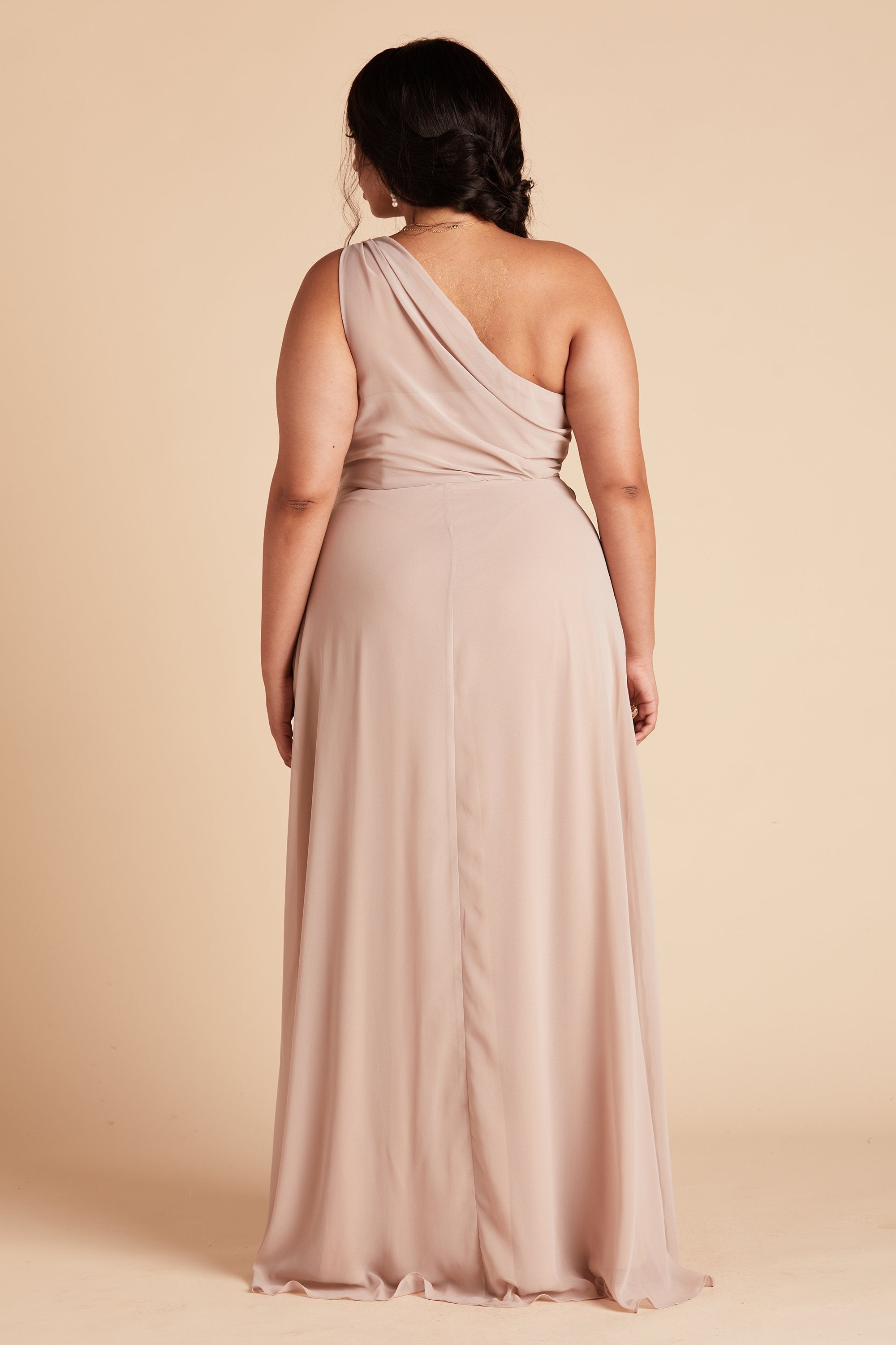 Back view of the Kira Dress Curve in taupe chiffon shows a full-figured model with a medium skin tone. The back of the dress has sheer chiffon fabric which creates a pleated Grecian one-shoulder neckline and bodice. The dress flows smoothly to the floor. 