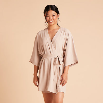 Front view of the Karen Robe in taupe shows a slender model with a medium skin tone wearing a short kimono-style robe that wraps across the chest and ties to one side at the waist. Wide, three quarter sleeves flow from the shoulder to the middle of the lower arm. The robe fits loosely over the body and is mid-thigh length. 